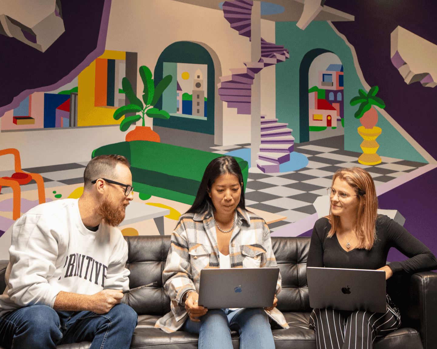 Employés m&h collaborant sur des ordinateurs portables, assis devant une fresque murale colorée au bureau.